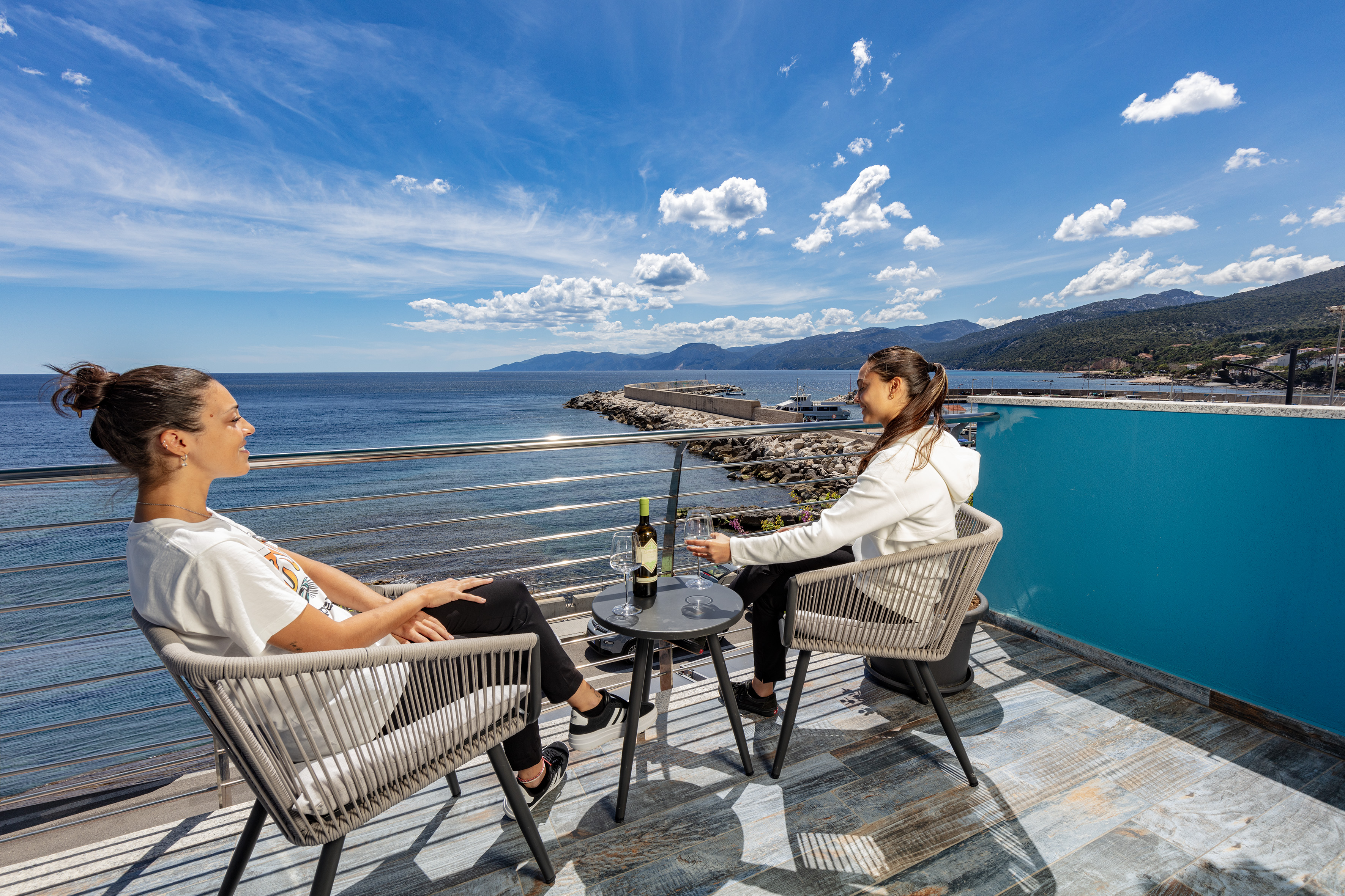 Due donne su sedie intrecciate con vino su un tavolino, terrazzo affacciato sul porticciolo e sui monti, La Casa del Pescatore