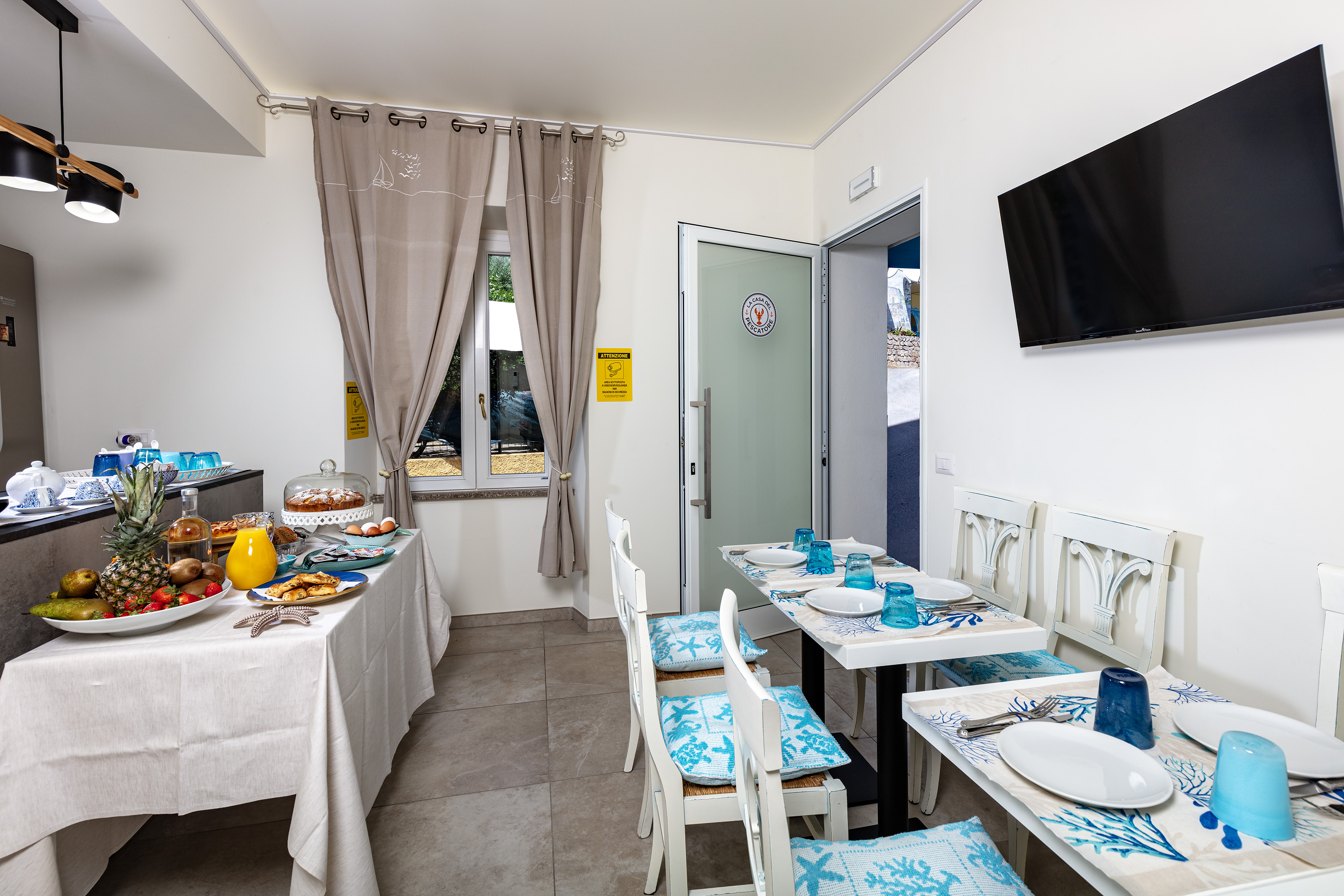 Sala colazione de La Casa del Pescatore con buffet di frutta e torta, sedie bianche e tovagliette a coralli azzurri