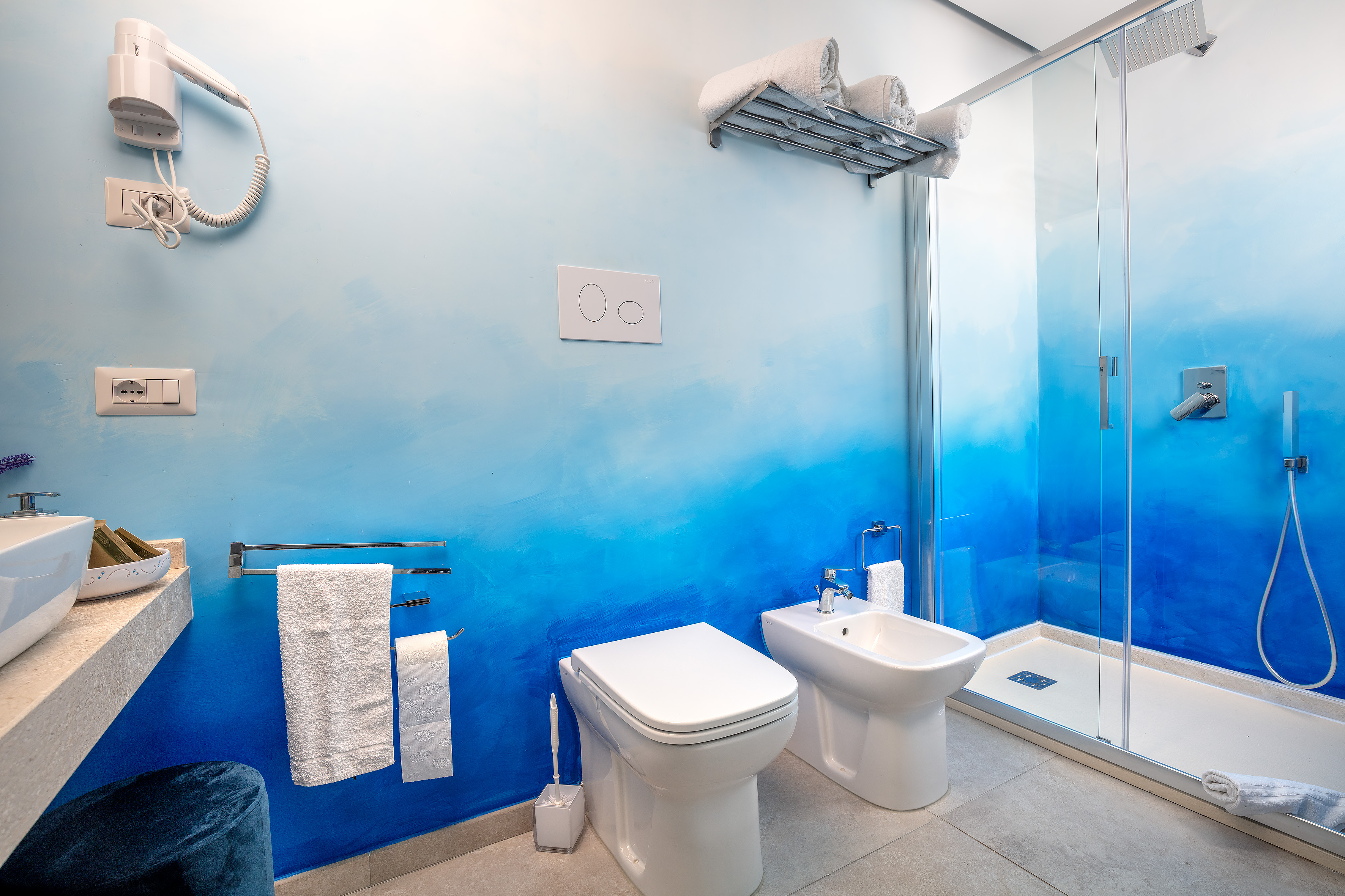 Bagno de La Casa del Pescatore con pareti sfumate dal celeste al blu, doccia in vetro, sanitari bianchi e asciugacapelli